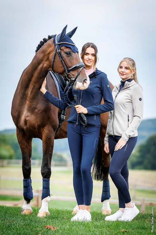 Two women in equestrian attire standing next to a horse in an outdoor setting.