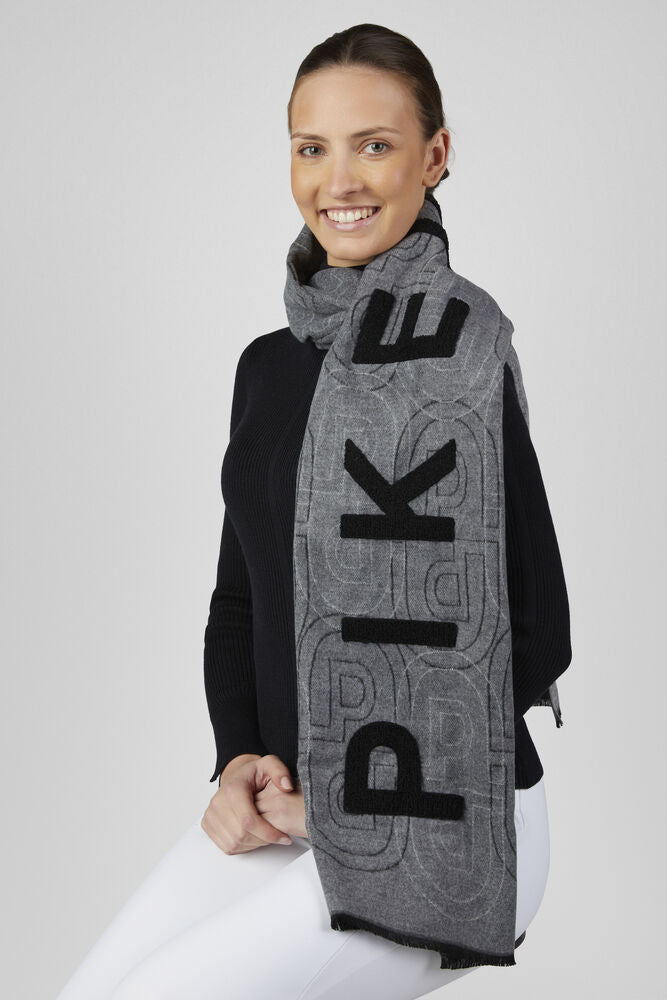 A woman wearing a gray knit scarf with black 'PIKEUR' lettering and a subtle geometric pattern, styled over a black top and white pants against a light gray studio background.