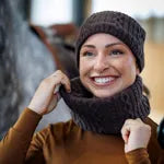 A smiling woman wearing a dark brown knit beanie and matching knit neck warmer, holding the neck warmer in place. She is wearing a brown top, with a blurred outdoor background.