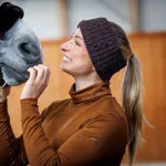A woman wearing a brown long-sleeve top and a dark knit beanie holds a gray horse close to her face in an indoor setting with warm wood walls.
