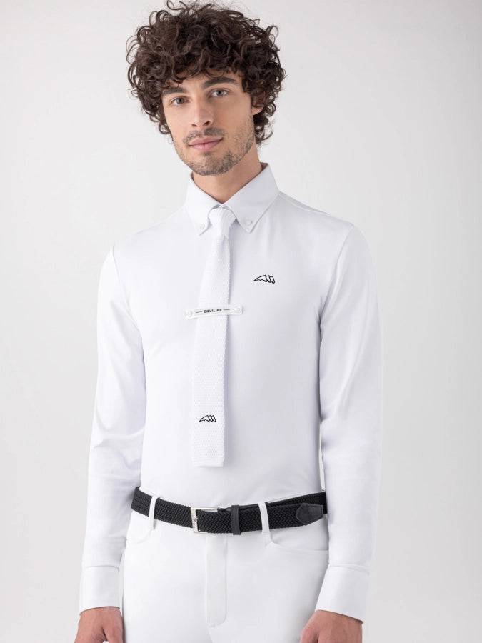 A man with curly dark hair wearing a white long-sleeve dress shirt, white tie, white trousers, and a black belt, photographed against a light gray studio background.