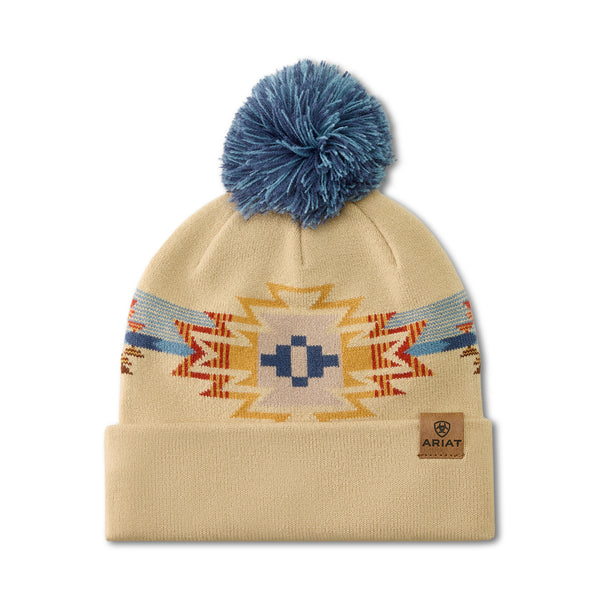 A beige knit beanie with a blue pom-pom on top, featuring a Southwestern-style geometric pattern in blue, orange, and yellow. A small brown leather patch on the cuff displays the ARIAT brand logo.