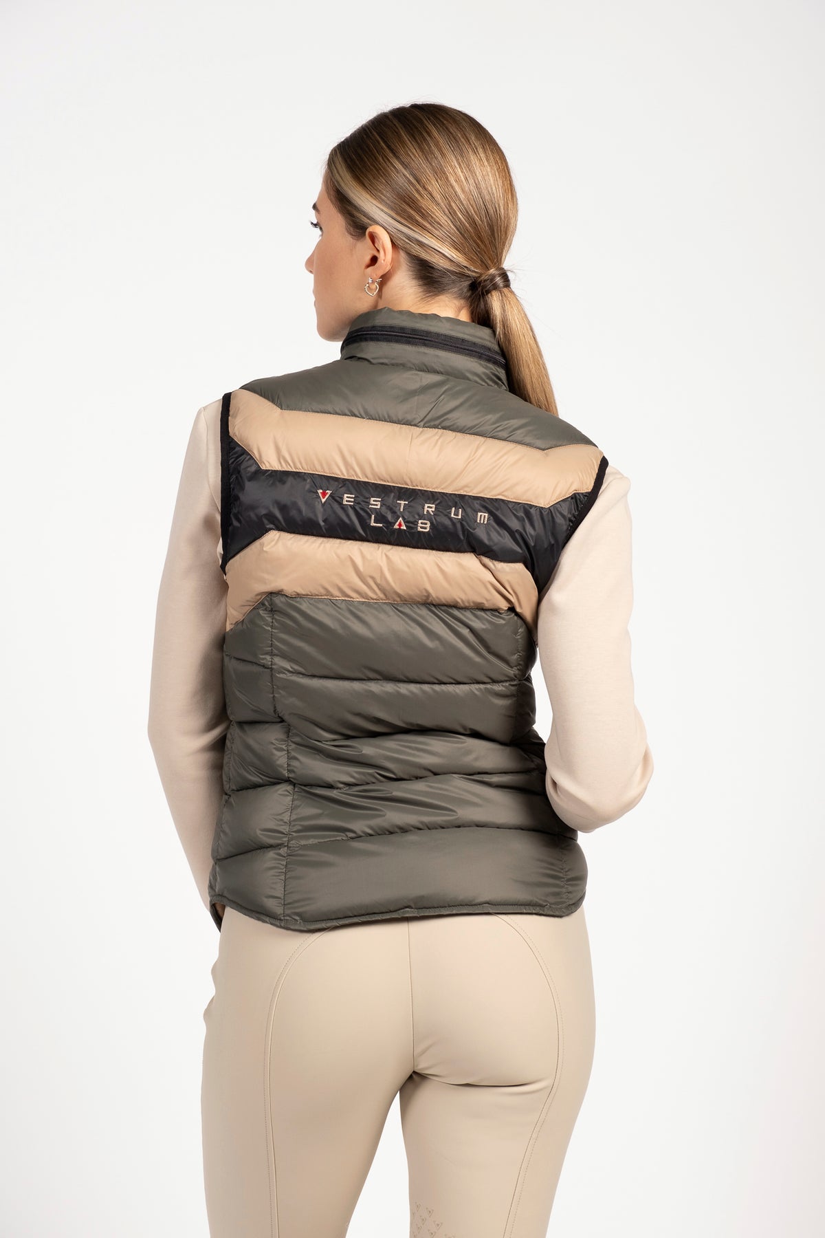 A woman shown from the back wearing a puffer vest with horizontal color-blocked stripes in dark olive green, beige, and black. The brand name 'VESTRUM LAB' is printed across the black stripe.