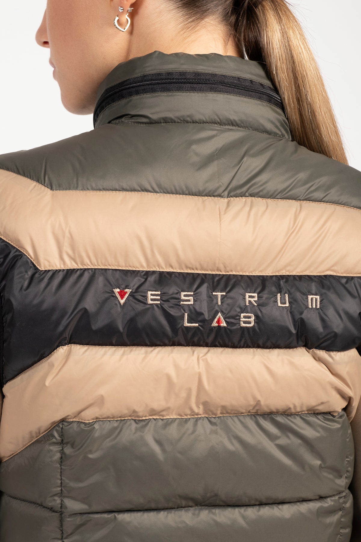 Close-up of a person wearing a puffer jacket with horizontal color-blocked stripes in olive green, beige, and black. The brand name 'VESTRUM LAB' is printed across the black stripe with a small red heart logo.