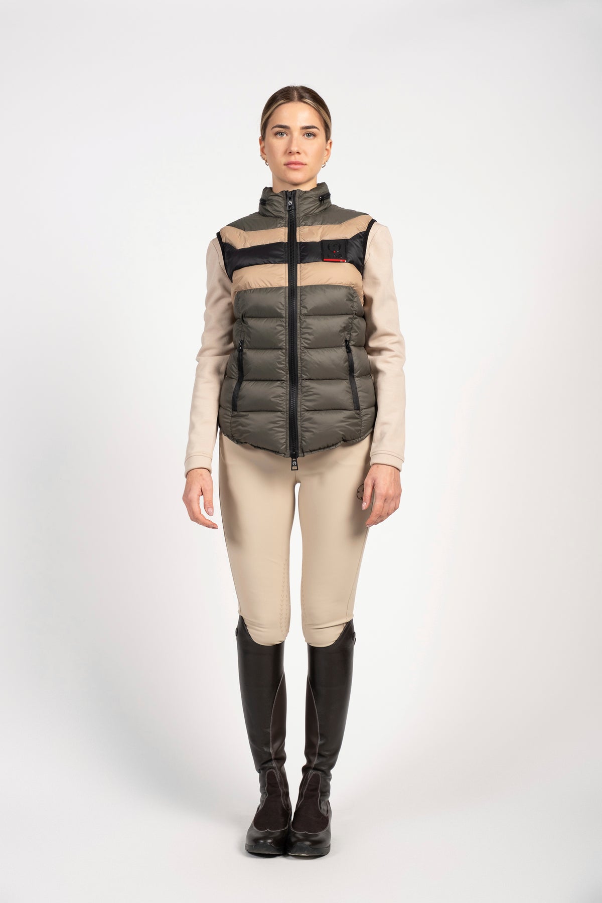 A woman stands against a light gray studio background wearing a sleeveless puffer vest with horizontal beige, black, and brown stripes over a beige long-sleeve top, beige riding breeches, and tall black riding boots.