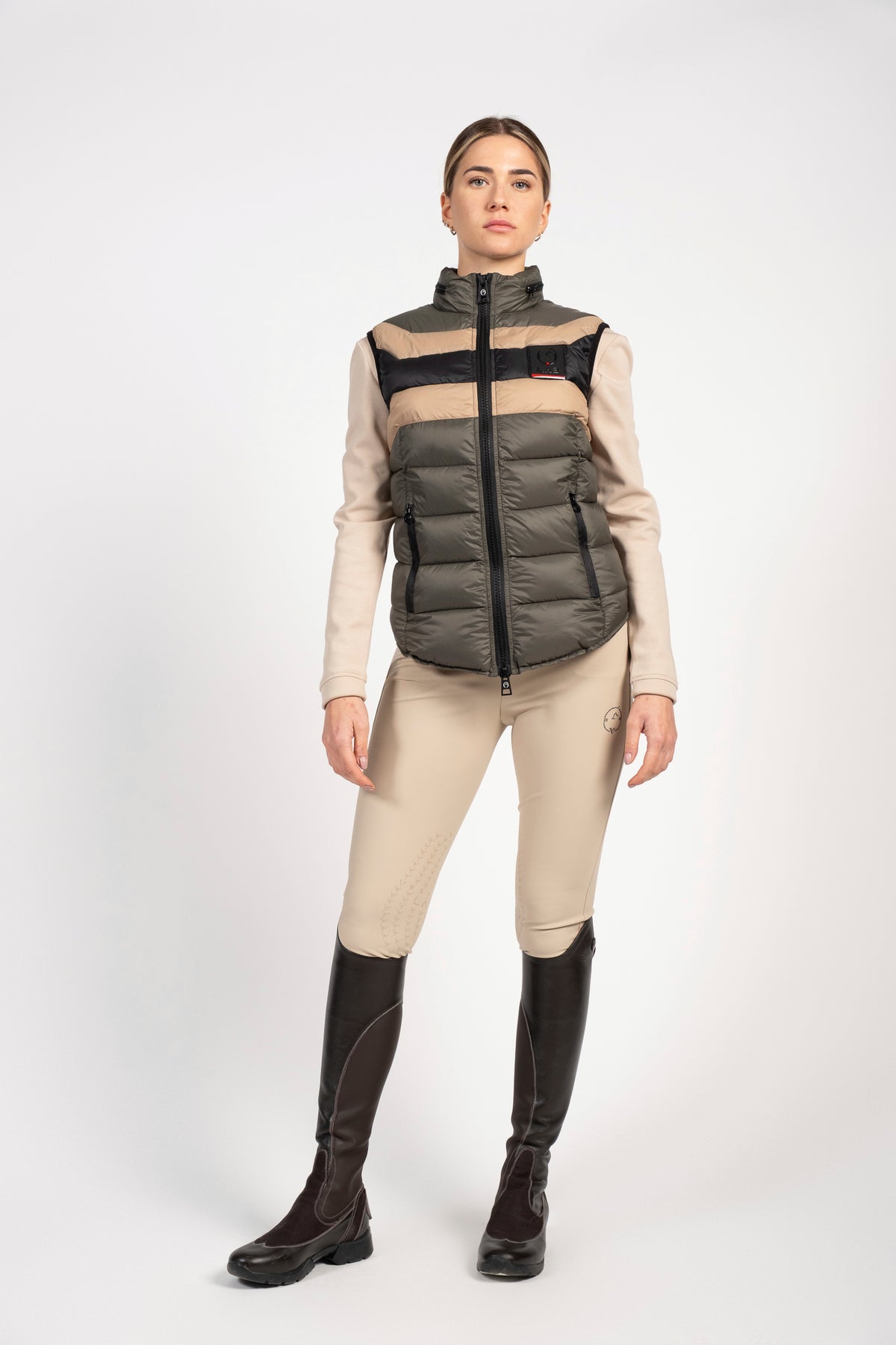 A woman modeling a color-blocked equestrian puffer vest with beige and dark gray horizontal stripes, paired with beige riding breeches and dark brown riding boots, against a light gray studio background.