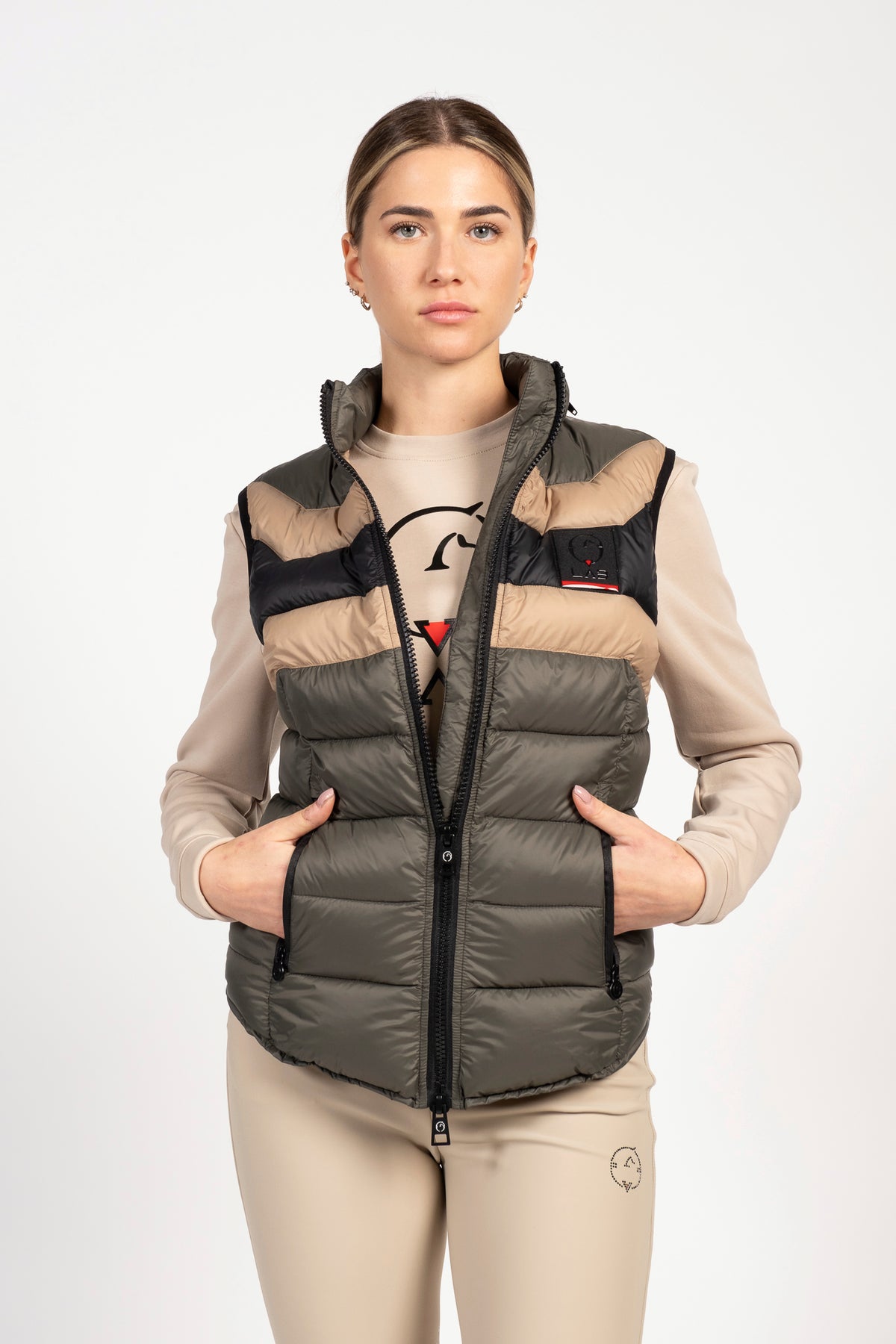 A woman modeling a sleeveless puffer vest with horizontal quilting. The vest features a color-blocked design with dark olive green, beige, and black panels, a black front zipper, and a small red and white logo on the left chest.