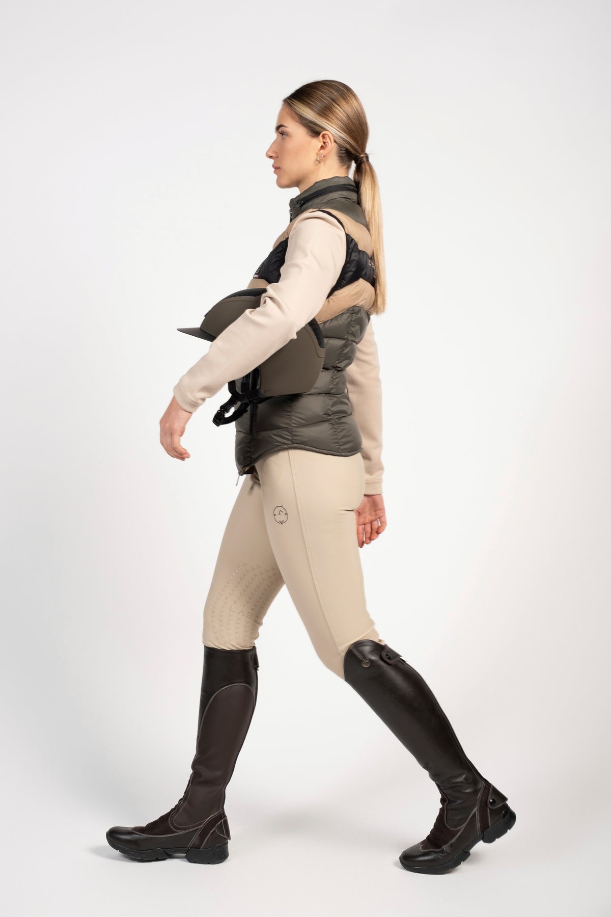 A woman in side profile wearing a dark gray quilted equestrian vest over a beige long-sleeve shirt, beige riding breeches, and black riding boots, against a plain white studio background.