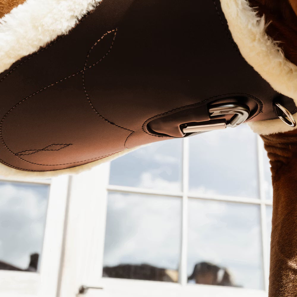 Close-up of a brown leather girth with fur lining, blurred window background