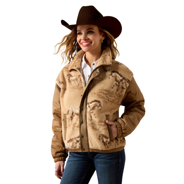 A woman wearing a brown cowboy hat and a sherpa fleece jacket with a brown and tan western horse-patterned design, paired with blue jeans, against a solid white background.