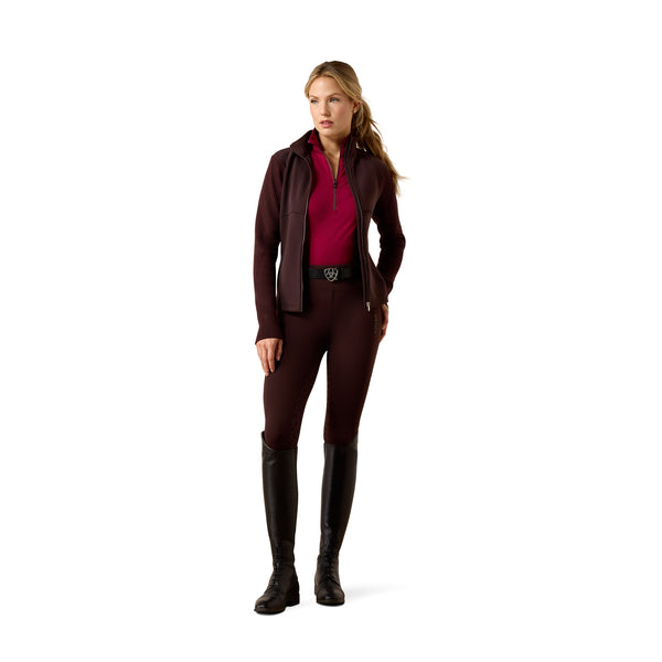 A woman modeling a dark burgundy equestrian riding jacket, matching riding pants, a magenta inner top, and tall black riding boots against a solid white background.