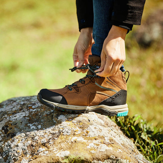 Person tying the laces of a brown hiking boot on a rock with a natural background