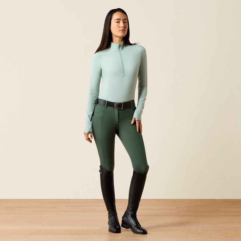 Woman wearing a light blue top and green riding pants with black boots on a beige background