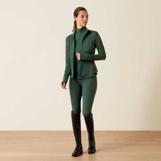 Woman wearing a green equestrian outfit with a jacket, leggings, and boots on a beige background