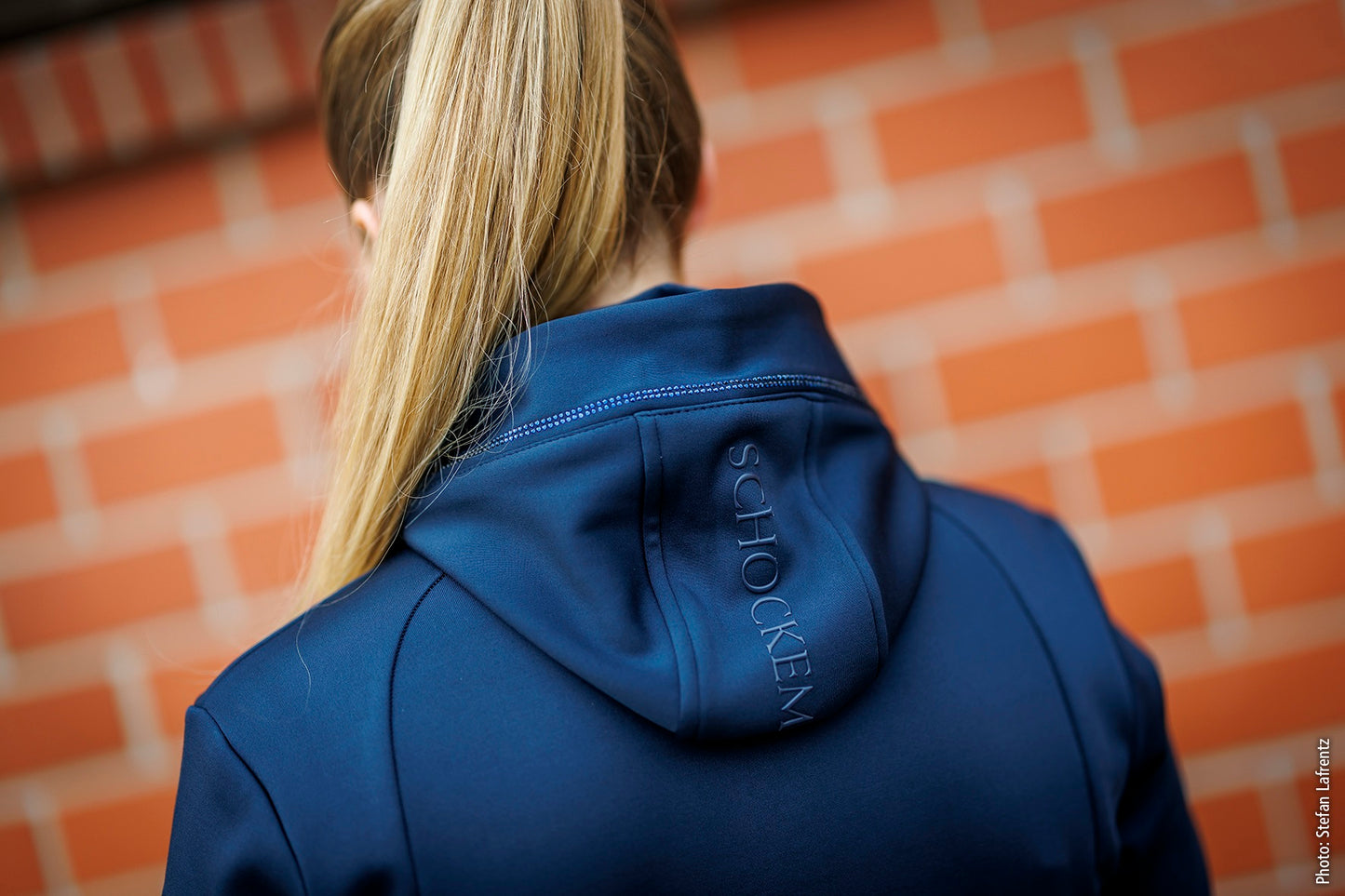 A woman viewed from behind wearing a dark blue hooded jacket with 'SCHOCKEMOHLE' embroidered vertically on the back, standing in front of a blurred orange brick wall.