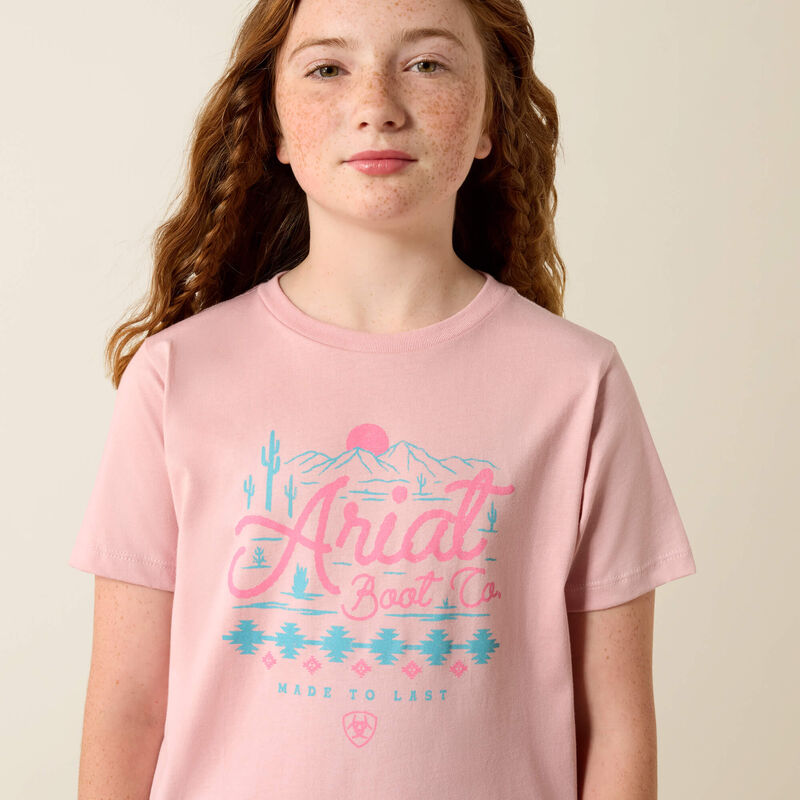 Young girl wearing a pink t-shirt with 'Ariat Boot Co.' graphic on a beige background