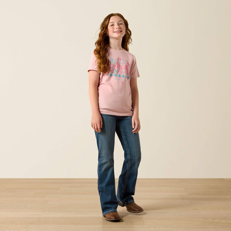 Young girl wearing a pink t-shirt and blue jeans standing on a wooden floor with a plain background