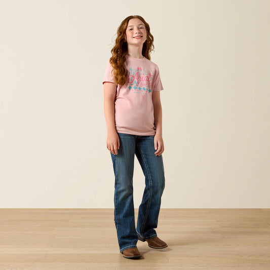 Young girl wearing a pink t-shirt and blue jeans standing on a wooden floor with a plain background