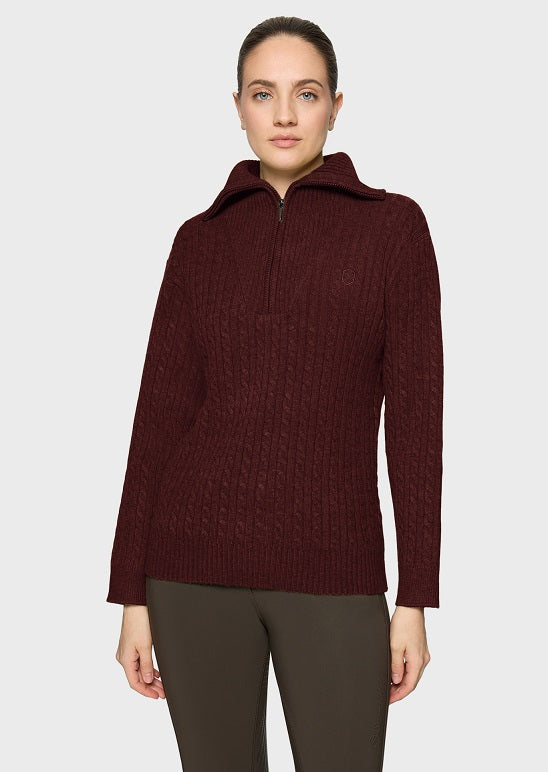 A woman modeling a dark burgundy ribbed knit half-zip pullover sweater with a stand-up collar, paired with dark olive pants, against a light gray studio background.