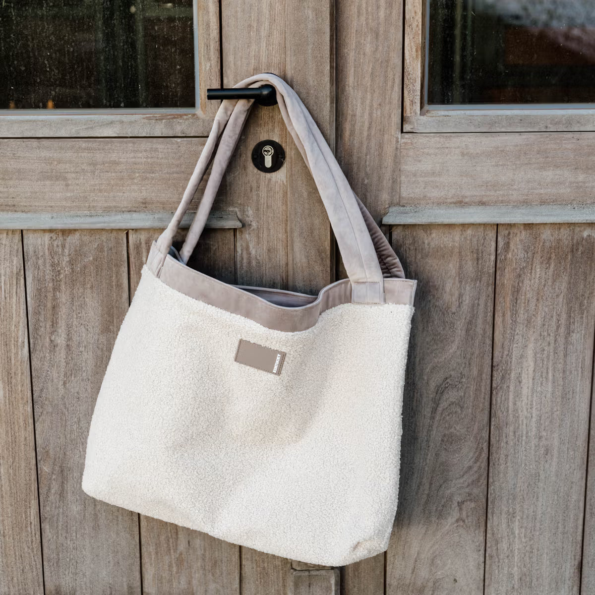 Beige tote bag with a brand label hanging on a wooden door.