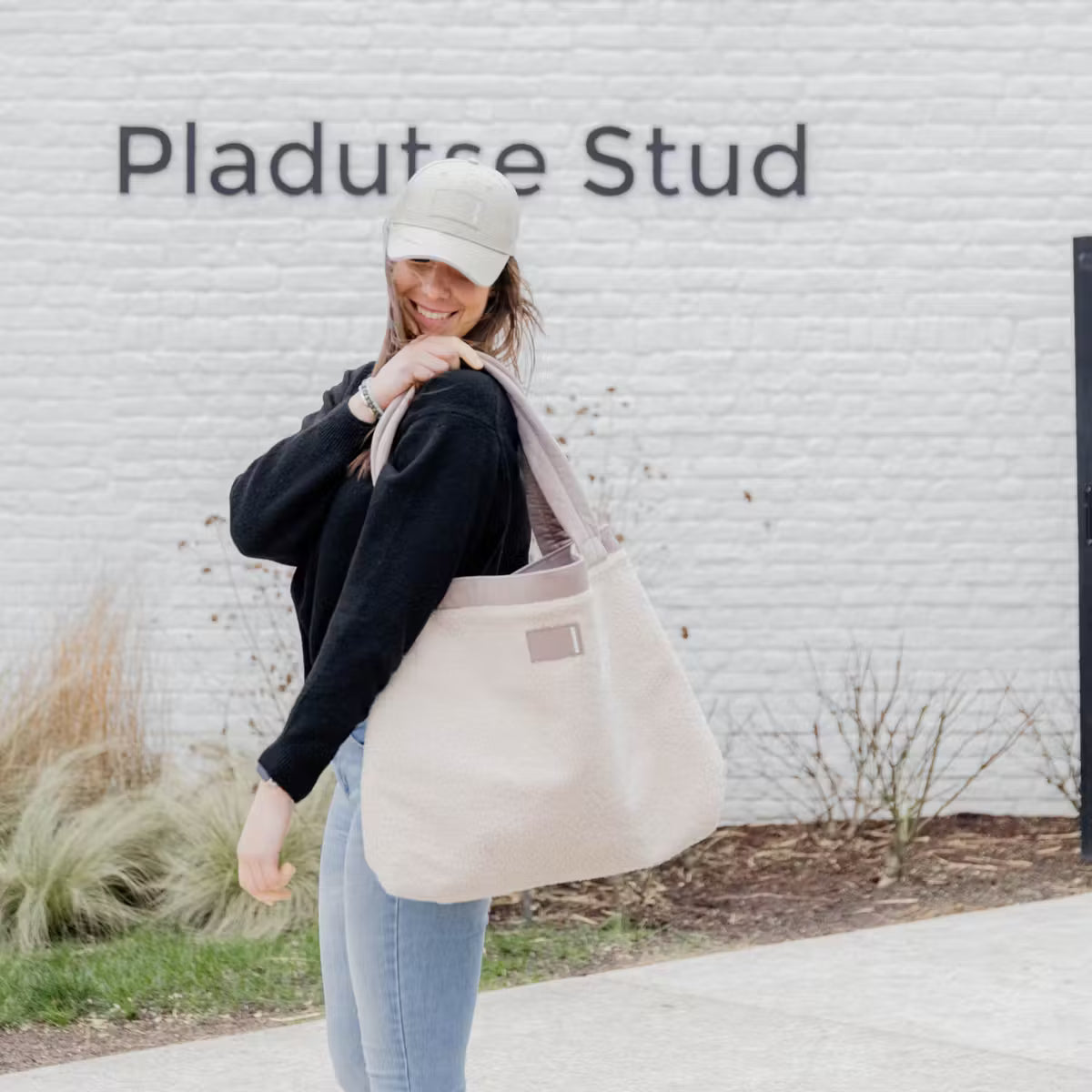 Woman holding a beige tote bag in front of a white wall with 'Pladute Stud' text.
