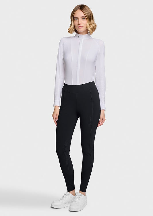A woman modeling a white long-sleeve mock-neck athletic top paired with black leggings and white sneakers, standing against a light gray studio background.