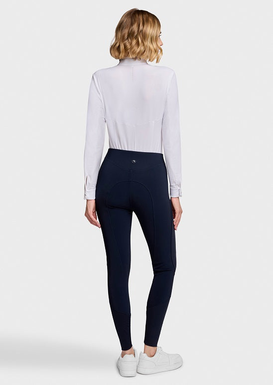 A woman shown from the back wearing a white long-sleeve mock-neck top, navy blue riding breeches, and white sneakers against a light gray studio background.
