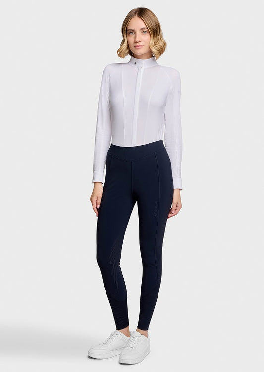 A woman modeling a white long-sleeve mock-neck athletic top paired with black leggings and white sneakers, standing against a light gray studio background.