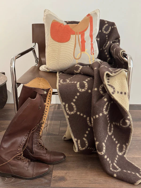 Brown and camel horse shoe blanket on a chair with a cushion depicting a saddle and a pair of brown riding boots next to it.