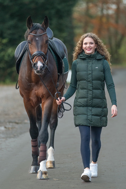 A woman walking alongside a saddled brown horse on a paved path, wearing a dark green puffer coat, dark leggings, and white sneakers.