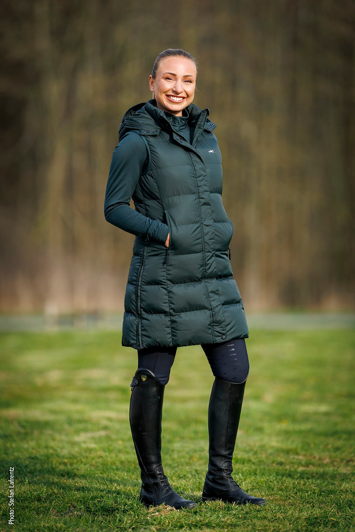 A woman standing outdoors on grass wearing a dark green hooded puffer vest over a matching long-sleeve top, with black riding boots and dark leggings, against a blurred wooded background.