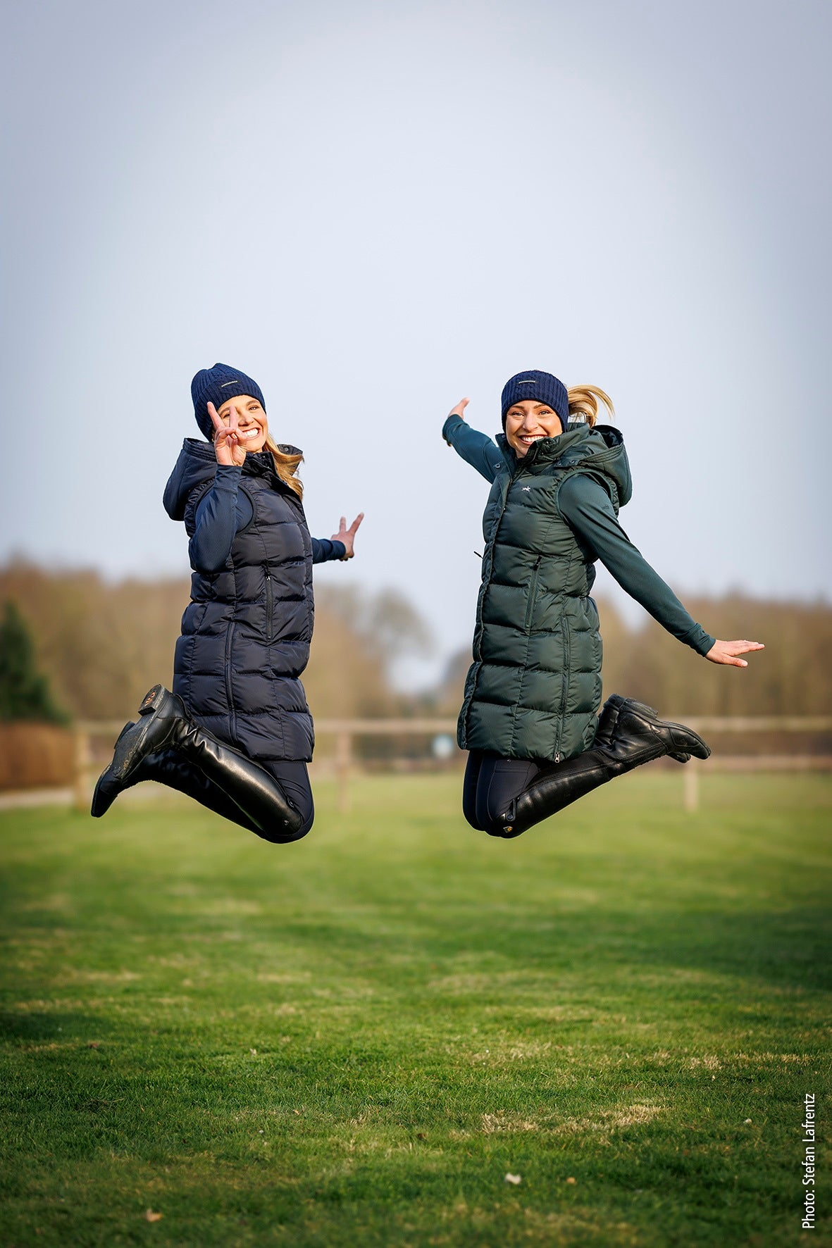 Two women jumping in the air outdoors on a grassy field, wearing long puffer coats and beanies. One coat is dark navy blue and the other is dark green, photographed under a pale sky.