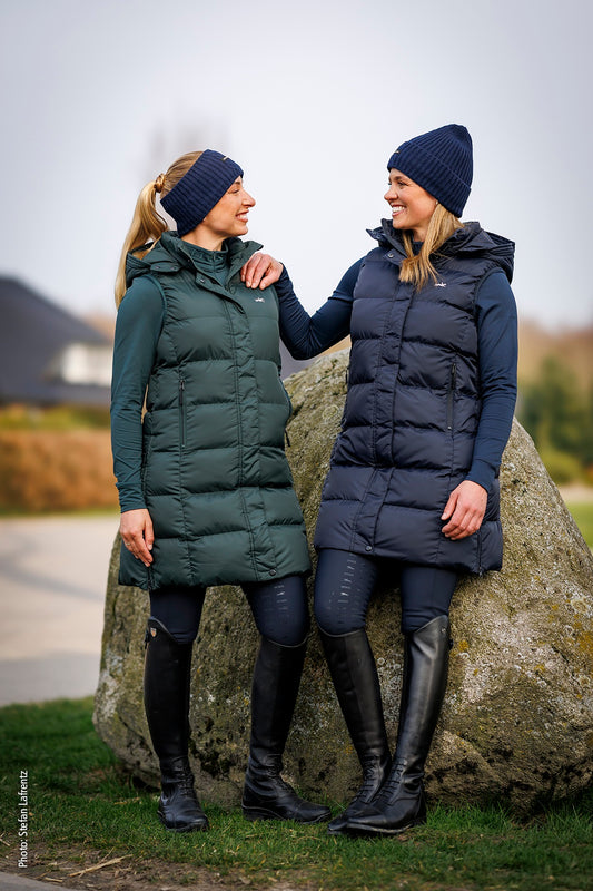 Two women standing outdoors next to a large rock, wearing long puffer vests in dark green and navy blue, along with matching knit beanies and tall black boots.