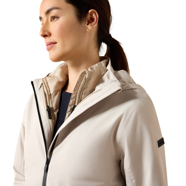 A woman in profile wearing a light beige technical outdoor jacket with a high collar and black zipper details, set against a solid white background.