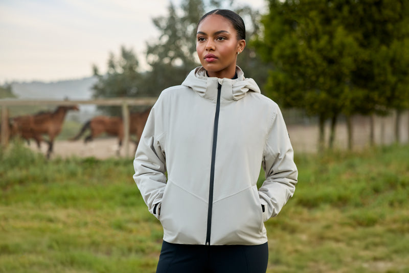 A woman wearing a light gray hooded zip-up jacket stands outdoors in a grassy field with trees and horses blurred in the background.