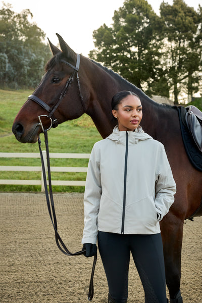 A woman wearing a light gray zip-up equestrian jacket and black riding tights stands in an outdoor horse arena next to a brown horse.