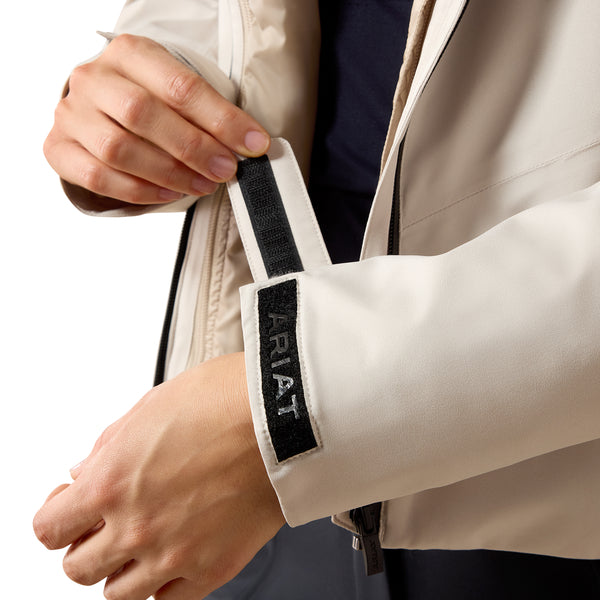 Close-up of a person wearing a light beige jacket, pulling open the front zipper while showing a black Velcro strap on the sleeve with the brand name ARIAT embroidered in white.