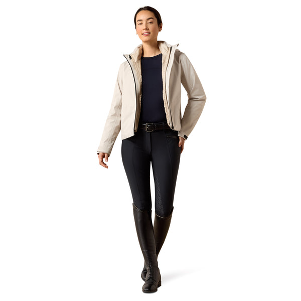 A woman modeling a light beige hooded jacket over a navy top, black riding pants, and black riding boots against a plain white background.
