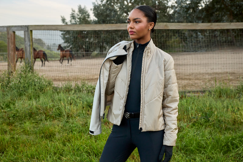 A woman stands outdoors in a grassy field wearing a light beige quilted jacket over a black top and black pants, with horses visible behind a fence in the background.