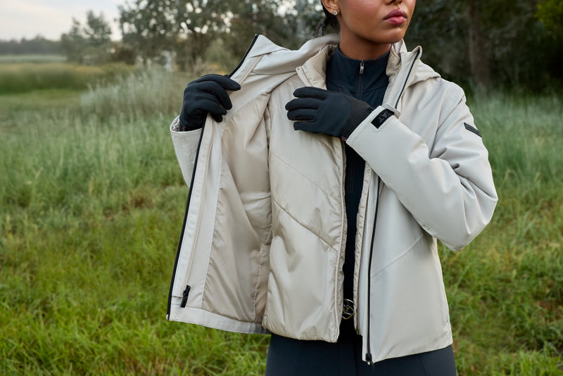 A woman outdoors in a grassy field wearing a light beige puffer jacket over a black top and black gloves, holding the jacket open to show the interior.