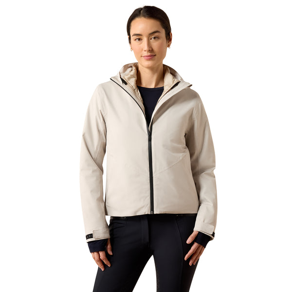A woman modeling a light beige hooded jacket with a black front zipper, worn over a dark navy top and black pants, against a plain white background.