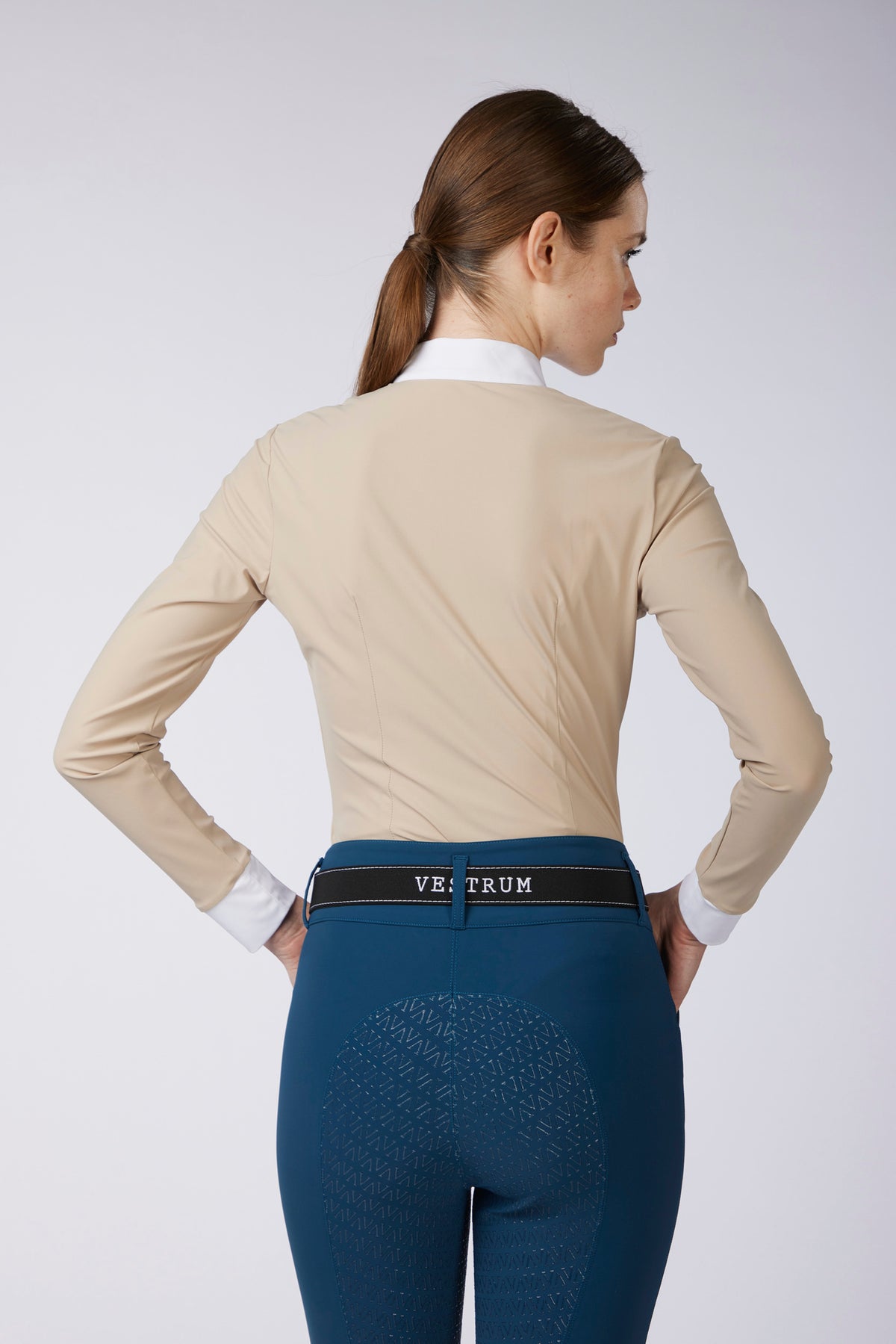 A woman shown from the back wearing a beige long-sleeve equestrian shirt, a white collared undershirt, a black riding belt with 'VESTRUM' branding, and dark teal riding breeches against a light gray studio background.