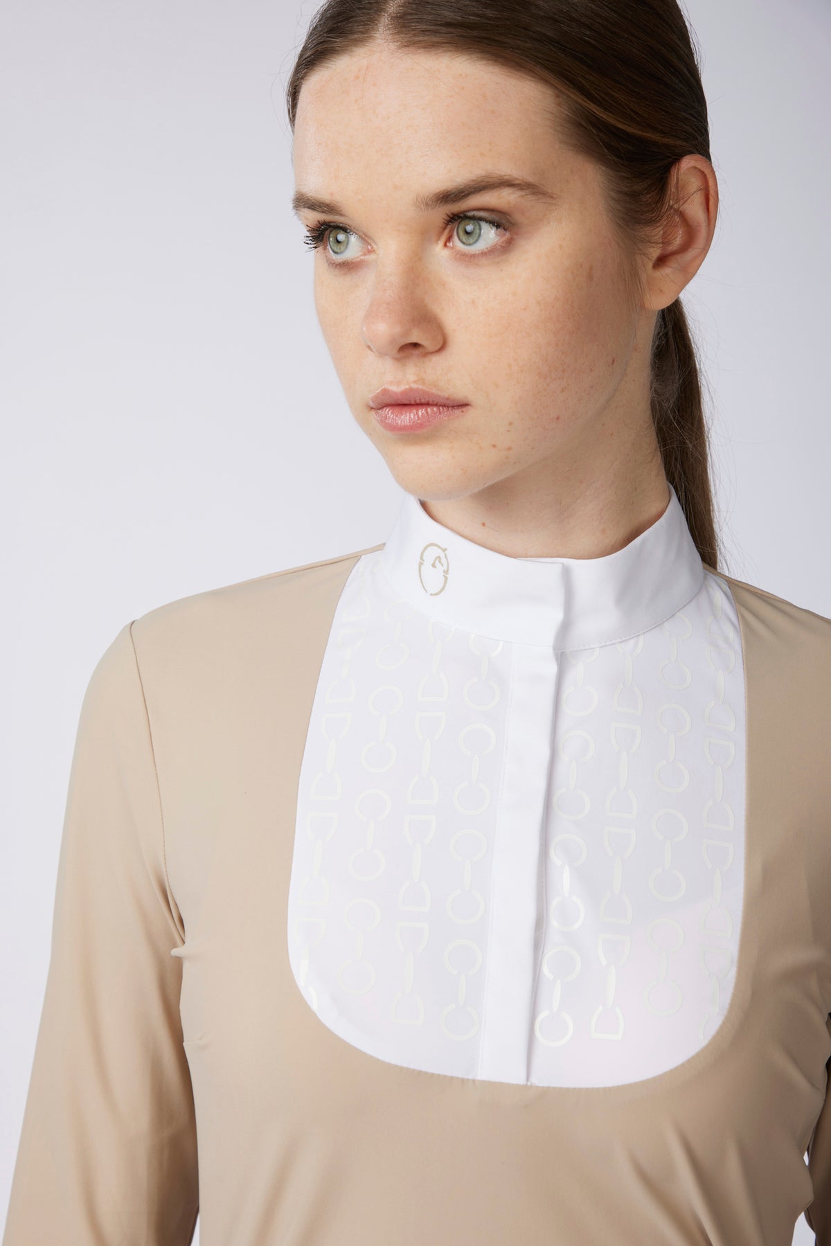 A close-up of a woman wearing a beige long-sleeve equestrian shirt with a white neck bib collar featuring a subtle embossed pattern.