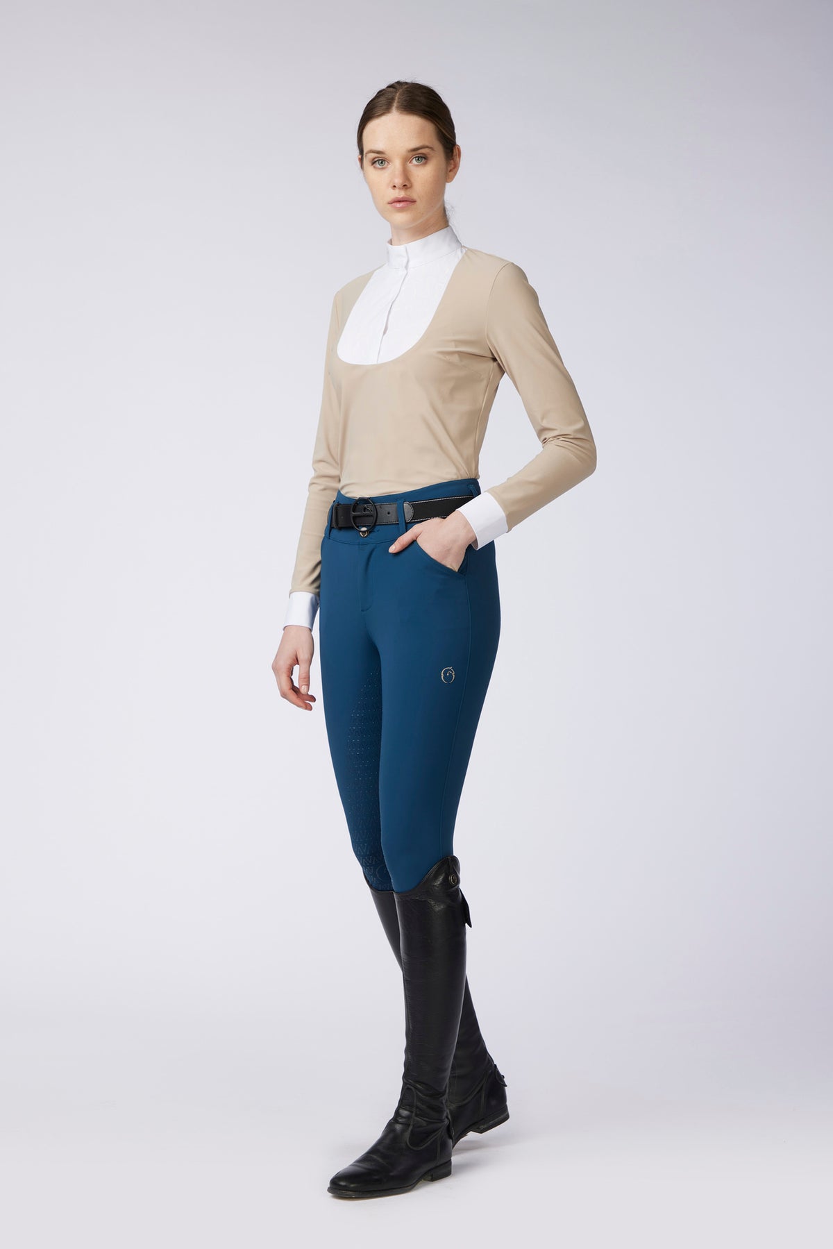 A woman modeling a beige long-sleeve riding shirt with a white collar, navy blue riding breeches, and black riding boots against a light gray studio background.