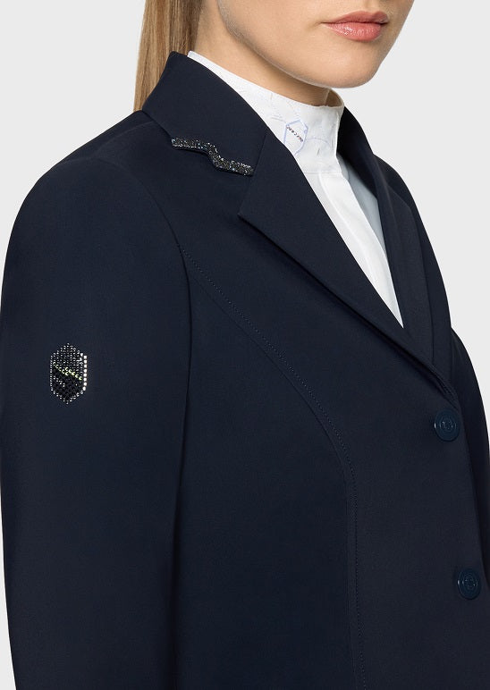 Close-up of a model wearing a dark navy blue tailored blazer over a white high-neck top. The blazer features decorative silver embellishments on the lapel and a small geometric emblem on the sleeve.