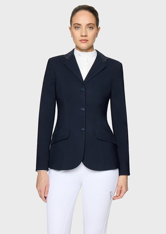 A woman modeling a dark navy equestrian blazer over a white high-neck shirt and white riding breeches, photographed against a light gray studio background.