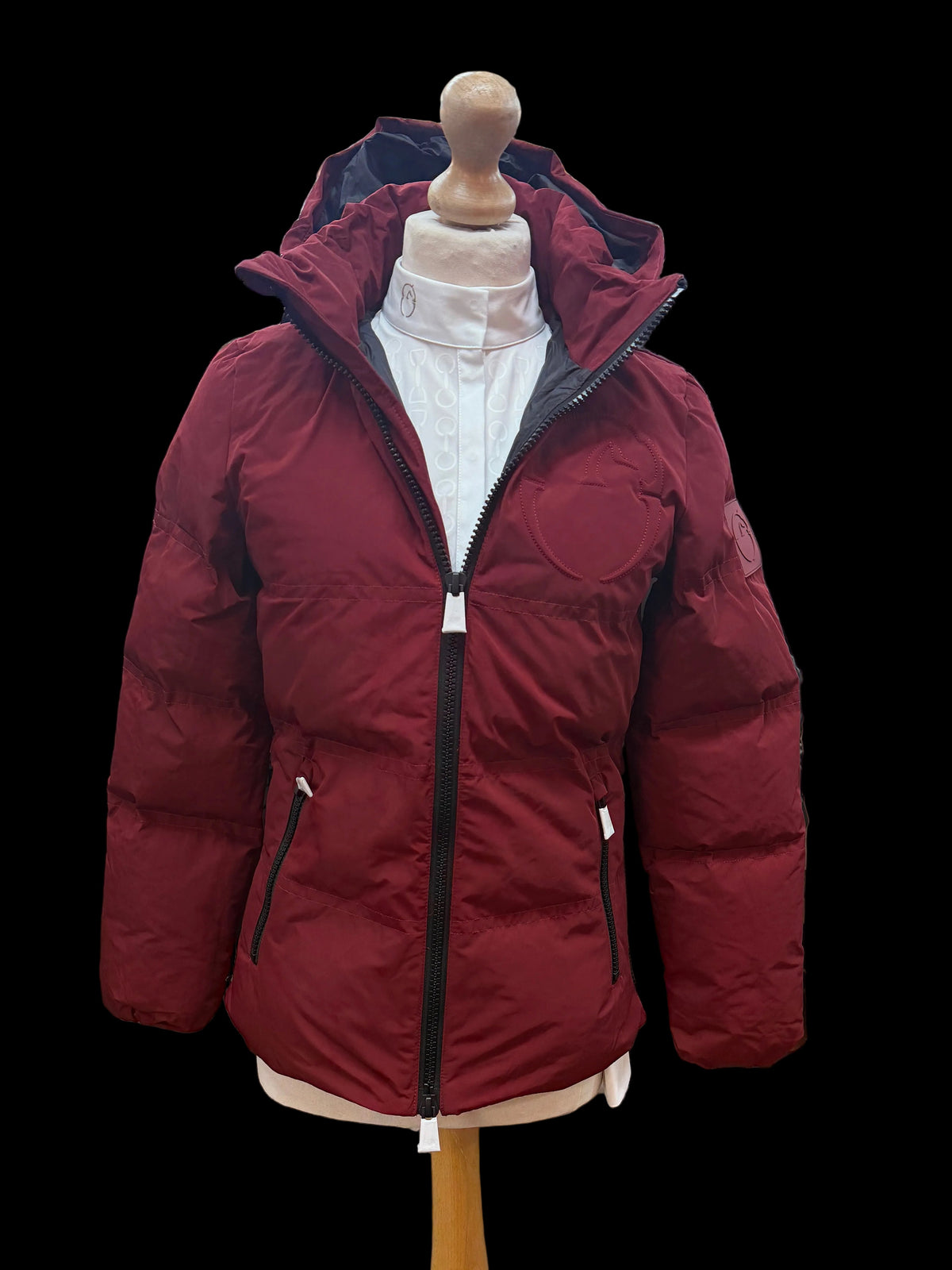 A red puffer jacket displayed on a wooden dress form mannequin inside a clothing store. The jacket features a high hood, a black front zipper, and zippered hand pockets.