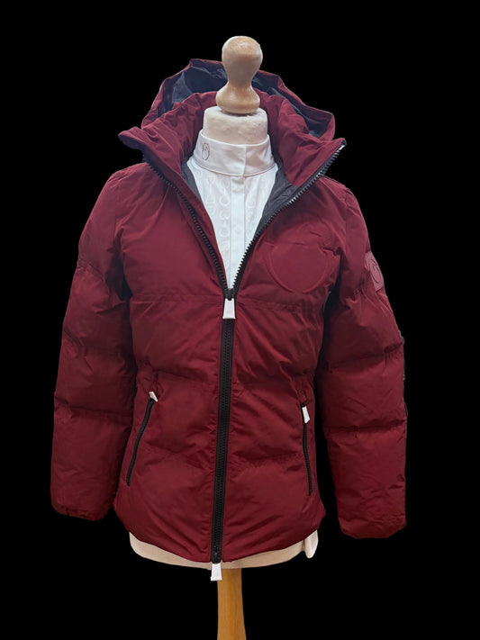 A red puffer jacket displayed on a wooden dress form mannequin inside a clothing store. The jacket features a high hood, a black front zipper, and zippered hand pockets.