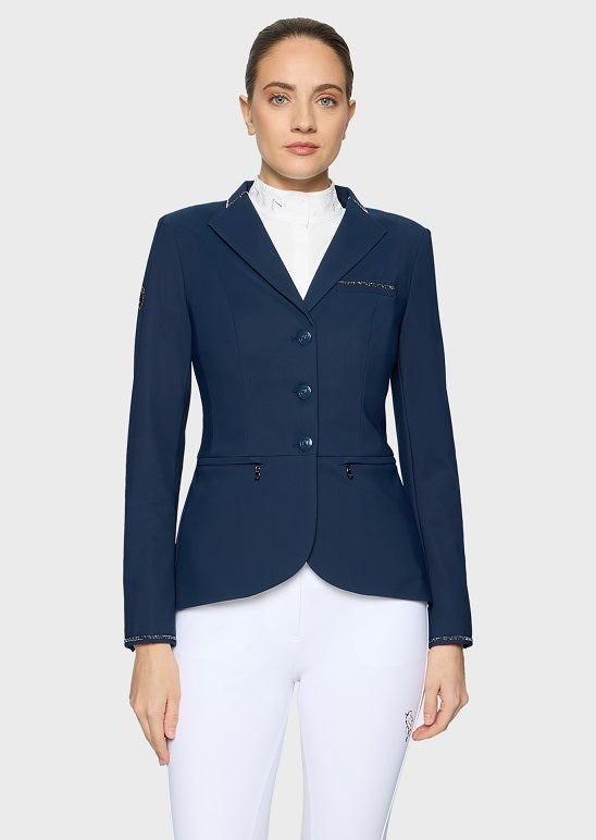 A woman modeling a navy blue equestrian-style blazer with white trim over a white high-neck shirt and white riding pants, against a light gray studio background.