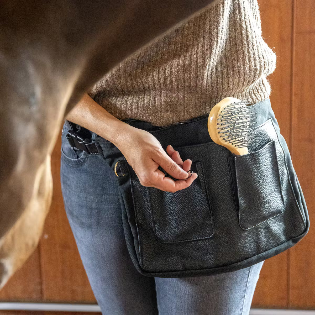 Person holding a black leather apron with a horse grooming brush in a stable setting